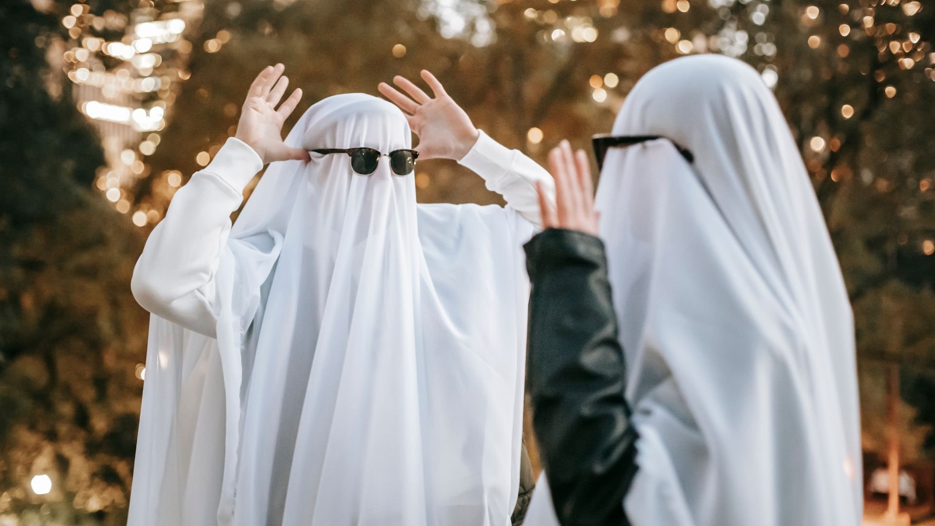 Two costumed ghosts make funny faces at each other while wearing sunglasses.