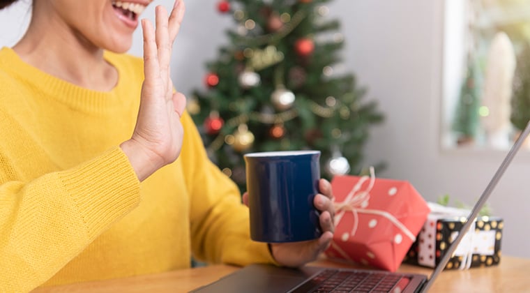 Woman waving at laptop camera with presents on desk and Christmas tree in background